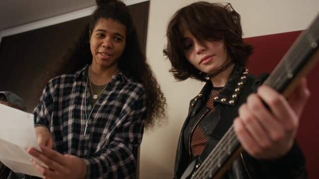 Multiethnic teenagers rehearsing rock music in studio, African American girl reading lyrics with Caucasian girl playing bass guitar. Perfect for youth culture and music education campaigns
