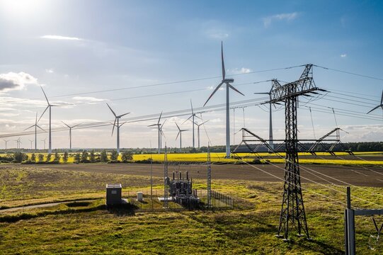 Large wind farm with electrical substation and transmission infrastructure in rural Germany. Modern wind turbines generate renewable electricity across agricultural fields under a dramatic sky,