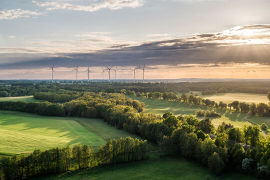 Aerial panorama of wind turbines above forests and green fields during sunset in Germany. Renewable energy infrastructure blends with the natural landscape under dramatic evening light.



