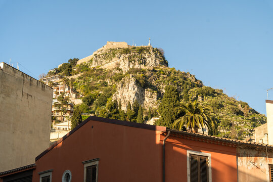 View of the historic Saracen Castle perched on the rocky cliffs of Monte Tauro above terracotta rooftops and lush greenery in Taormina, Sicily, Italy.