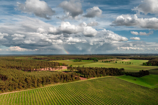 Aerial view of agricultural fields, forests and rolling countryside in Germany beneath dramatic storm clouds. A rainbow appears on the horizon while distant wind turbines symbolize renewable energy an
