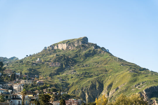 View of the lush green slopes and rocky peak of Monte Tauro overlooking residential buildings under a clear blue sky in Taormina, Sicily, Italy.