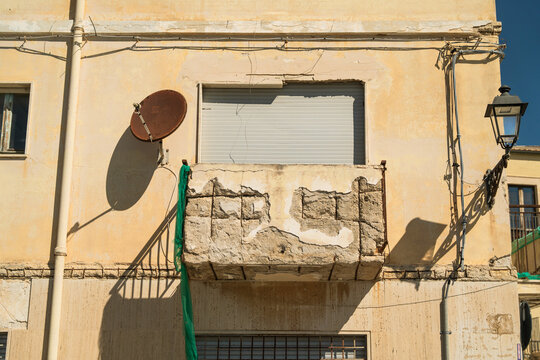 View of a weathered yellow building facade featuring a crumbling stone balcony with exposed rebar, a satellite dish, and a vintage street lamp in Syracuse, Sicily, Italy.