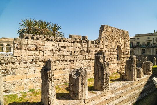 View of the ancient ruins of the Temple of Apollo with weathered stone walls and columns under a clear blue sky in Syracuse, Sicily, Italy.