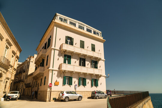 View of a historic white building with green shutters and stone balconies along a coastal road under a clear blue sky in Syracuse, Sicily, Italy.