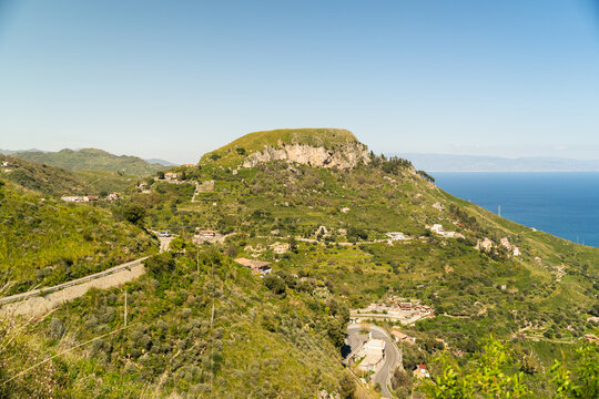View of the historic hilltop village nestled on a rocky outcrop with lush green slopes and the blue Mediterranean Sea in the distance Savoca, Sicily, Italy.