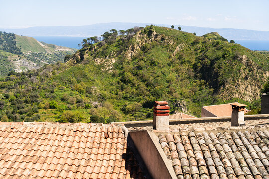 View of terracotta rooftops and chimneys overlooking lush green hills and the Mediterranean Sea under a clear sky in Savoca, Sicily, Italy.