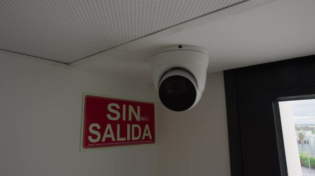 White dome surveillance camera fixed to a ceiling corner beside a window, with a slow camera push in toward the security lens monitoring the interior space