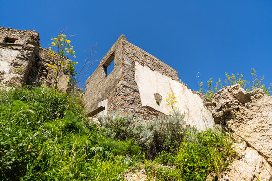View of ruined stone buildings and crumbling walls on a lush green hillside under a clear blue sky in Savoca, Sicily, Italy.