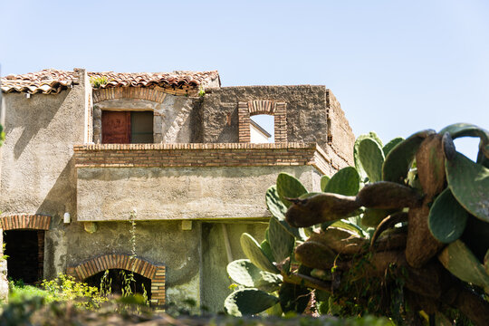 View of a rustic stone building with terracotta roof tiles and arched brick details next to a prickly pear cactus in Savoca, Sicily, Italy.