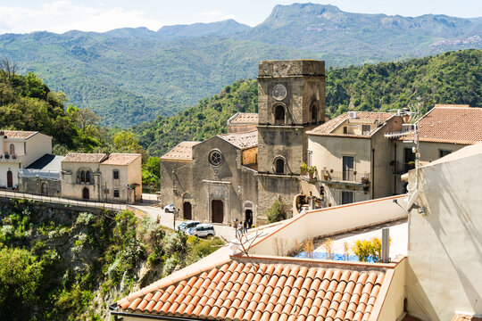 View of the historic San Nicolo church and traditional terracotta rooftops against a backdrop of lush green mountains in Savoca, Sicily, Italy.