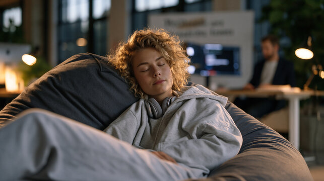 Young founder collapsed asleep on a beanbag in a tiny office while her business partner still codes at a standing desk behind her, a motivational whiteboard message half-erased, the imbalance of res