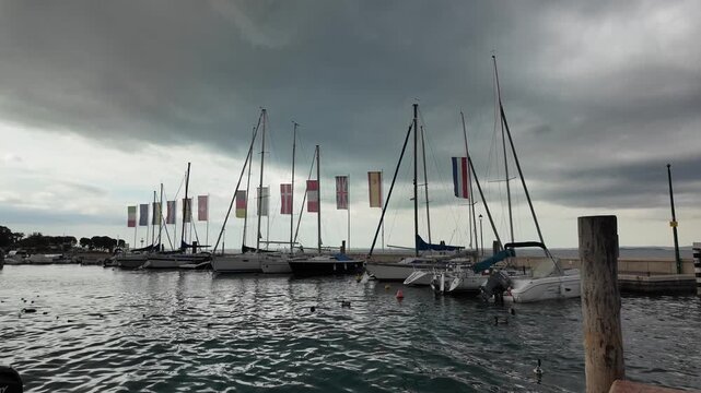 Bardolino, Italy - May 14, 2026 - Sailboats docked in a harbor under a dramatic cloudy sky with flags flying