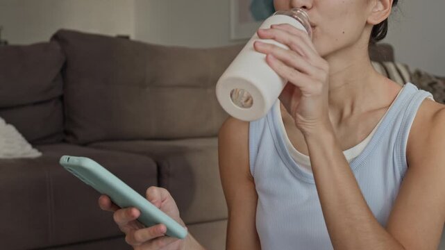 Young adult Asian woman holding smartphone and drinking water from bottle at home. Video perfect for wellness apps, dairy marketing, and healthy lifestyle campaigns