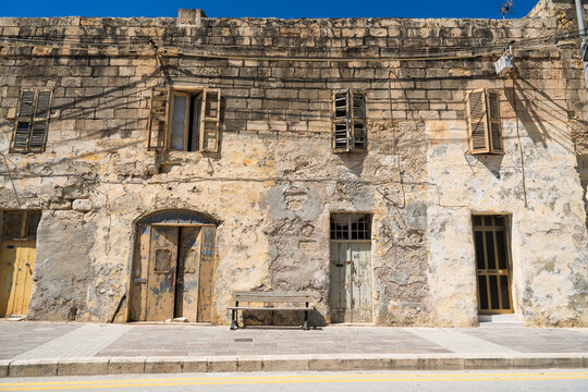 View of a traditional weathered limestone building facade with old wooden shutters and doors under a clear blue sky in Marsaskala, Southern Region, Malta.