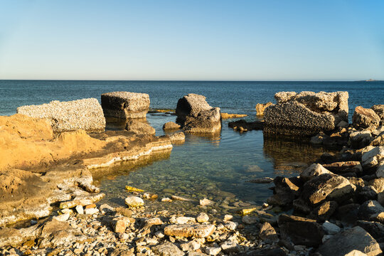 View of weathered concrete blocks and rocky shoreline along the clear Mediterranean sea under a bright blue sky in Marsaskala, Southern Region, Malta.