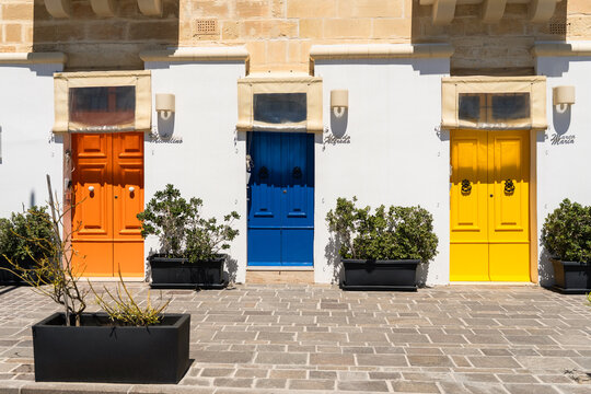 View of three vibrant orange, blue, and yellow wooden doors on a white facade with potted green plants and stone paving in Marsaskala, Southern Region, Malta.