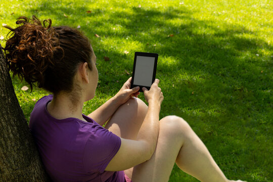 A woman leaning against a tree, reading an e-book, in a park on a sunny day