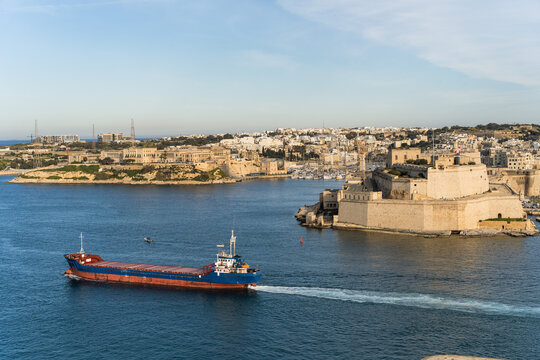 View of a cargo ship sailing through the Grand Harbour past the historic Fort Saint Angelo and the limestone architecture of the city Valletta, Port Region, Malta.