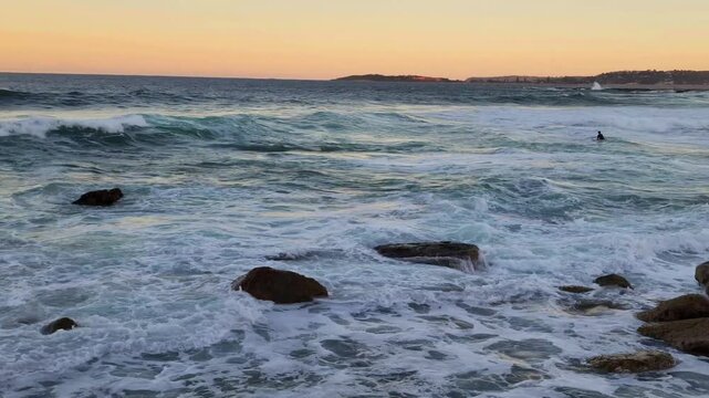 Sydney, Australia - The video illustrates a dramatic coastal scene on Sydney&rsquo;s Northern Beaches, where waves crash against shoreline rocks under warm sunset light. 