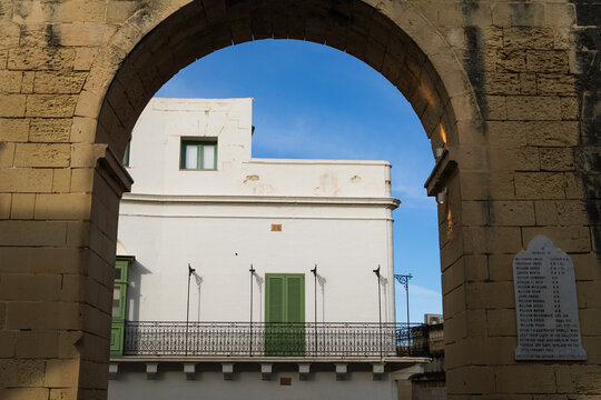 View of a traditional white building with green shutters and a wrought-iron balcony framed by a historic stone archway in Valletta, Port Region, Malta.