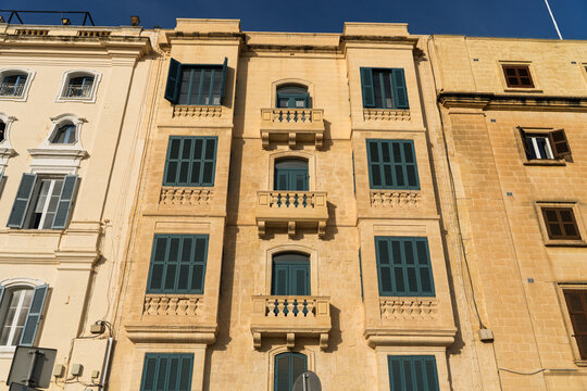 View of a traditional limestone building facade with green wooden shutters and stone balconies under a clear sky in Valletta, Port Region, Malta.