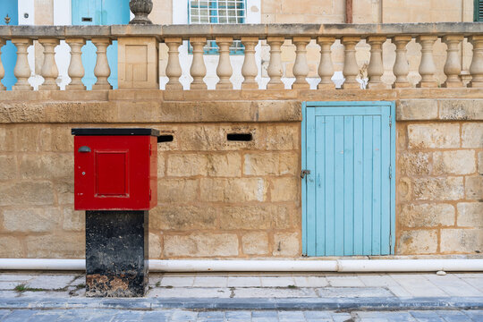 View of a red utility box and a small blue wooden door against a traditional limestone wall with a stone balustrade in Valletta, Port Region, Malta.