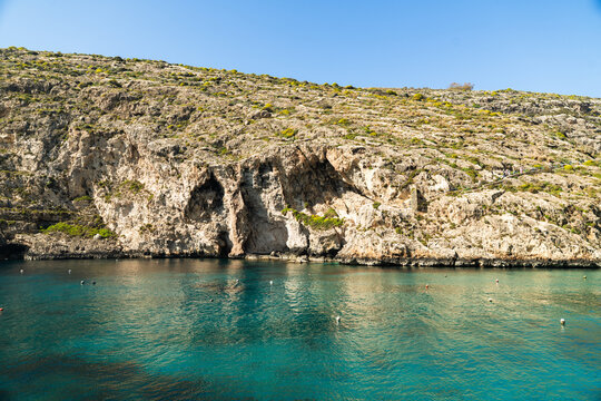 View of the turquoise waters of Xlendi Bay framed by rugged limestone cliffs under a clear blue sky in Zebbug, Gozo Region, Malta.