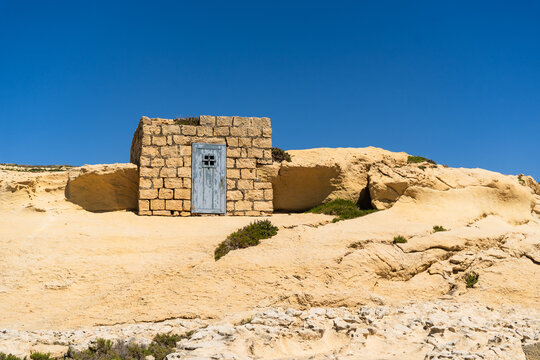 View of a small stone hut with a blue wooden door on the rocky cliffs of Dwejra Bay under a clear blue sky in Zebbug, Gozo Region, Malta.