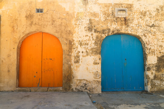 View of two arched wooden doors, one orange and one blue, set into a weathered limestone wall in Zebbug, Gozo Region, Malta.