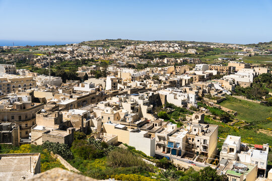 View of traditional limestone buildings and flat rooftops overlooking green fields and the Mediterranean Sea under a clear blue sky in Zebbug, Gozo Region, Malta.