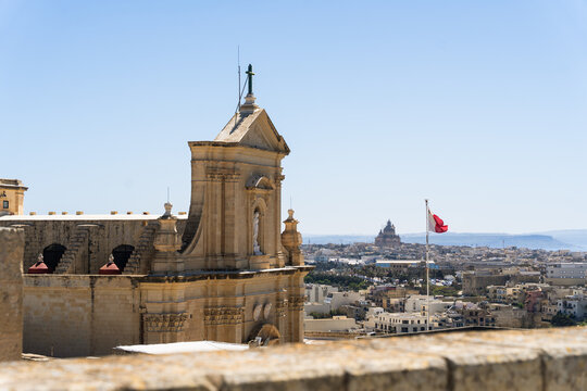 View of the historic Cathedral of the Assumption with its baroque facade and a Maltese flag waving against a clear blue sky Zebbug, Gozo Region, Malta.