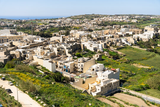 View of traditional limestone houses and narrow streets surrounded by lush green hills and fields under a clear blue sky in Zebbug, Gozo Region, Malta.