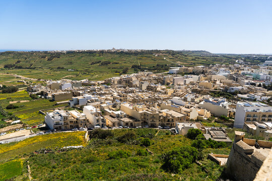 View of the town of Zebbug with light-colored stone houses and green agricultural fields under a clear blue sky in Zebbug, Gozo Region, Malta.