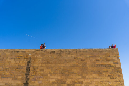 View of people sitting on top of a high limestone fortification wall under a clear blue sky in Zebbug, Gozo Region, Malta.