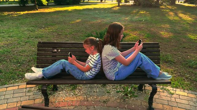Girls on a Park Bench with Smartphones, Siblings Sitting Back to Back, Children Absorbed in Gadgets, Outdoor Screen Time, Digital Generation, Park Bench Downtime, Kids with Mobile Phones, Modern