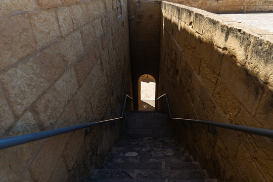 View of a narrow stone staircase descending between high walls toward an arched doorway in bright sunlight Zebbug, Gozo Region, Malta.