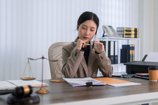 Asian female lawyer reviewing legal documents talking phone consultation professional office desk with scales justice gavel paperwork attorney workplace