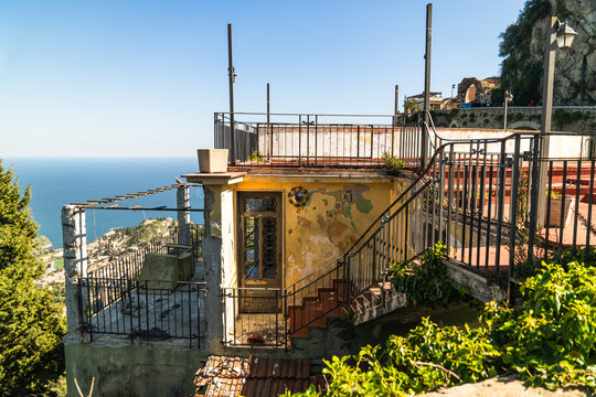 View of an old weathered building with peeling yellow paint and metal railings overlooking the blue Mediterranean Sea and the coastline from a high vantage point in Castelmola, Sicily, Italy.