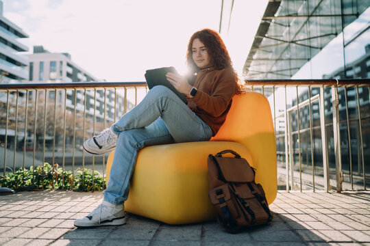 Young professional with backpack sitting in yellow chair using digital tablet in modern city. Remote office concept, digital nomad lifestyle, student life, commuting freelancer, urban environment.