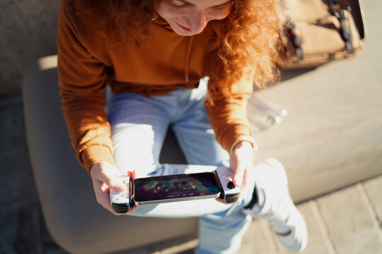 High angle of ginger model playing mobile video game with gamepad outdoors. Esports lifestyle, mobile application search, multimedia content, wireless technology, 5G network, leisure.