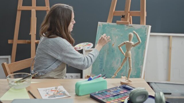 Young woman painter wearing apron holds palette and paints canvas with hand at easel in studio while smiling; creative joy.