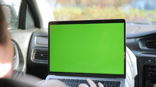 Person works on laptop with green screen inside car. Fingers type on keyboard, focused on task. View from behind, emphasizing work environment. Useful for remote work, travel