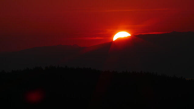 View of a vibrant red sunset with the sun dipping behind the Jura mountains and a silhouetted forest in Gex, Auvergne-Rhone-Alpes, France.