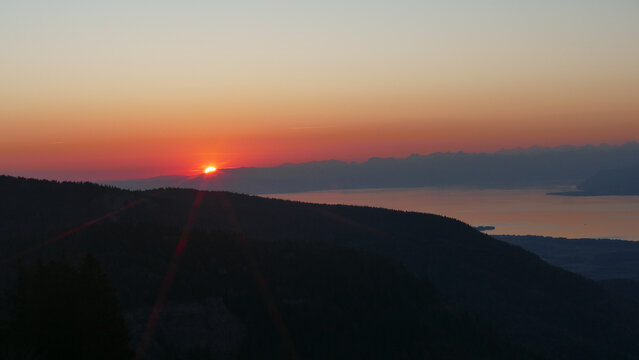 View of the sun setting over Lake Geneva with dark mountain silhouettes and a glowing orange sky in Gex, Auvergne-Rhone-Alpes, France.