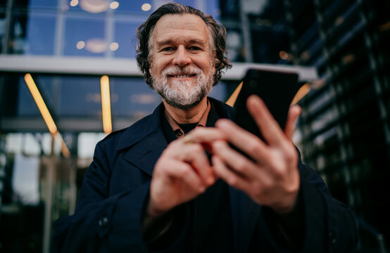 Portrait of cheerful senior businessman holding smartphone and looking at camera with joy, successful mature male professional using mobile device in contemporary city environment, copy space.
