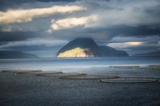 View of Koltur island with sunlight hitting its green slopes and circular fish farm pens in the sea under dramatic clouds Koltur, Streymoy, Faroe Islands.