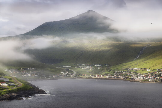 View of the coastal village of Sandavagur with its red-roofed church nestled at the base of a misty mountain peak by the bay in Sandavagur, Vagar, Faroe Islands.
