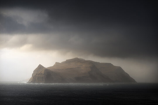View of Mykines island illuminated by a patch of sunlight under dramatic dark storm clouds over the North Atlantic Ocean Mykines, Vagar, Faroe Islands.