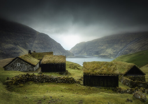 View of traditional turf-roofed houses and stone walls overlooking a lagoon under dramatic dark clouds in Saksun, Streymoy, Faroe Islands.